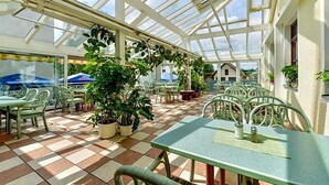Un patio cubierto con mesas y sillas, plantas en macetas y una estructura de techo de cristal.