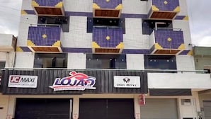 Un edificio de varios pisos con fachada de tejas, que cuenta con tres locales comerciales a nivel de calle con señalización visible para 'JC MAXI', 'LOJA' y 'PRO MAXI'.