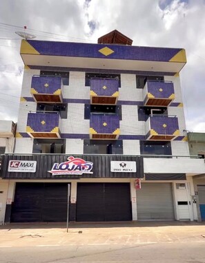 A multi-story building with a tiled facade, featuring three ground-level commercial spaces with visible signage for 'JC MAXI,' 'LOJA,' and 'PRO MAXI.'