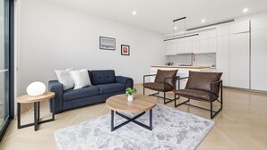 A modern living room with a sofa, two armchairs, a coffee table, and a small side table. There is a kitchen area with white cabinets and a countertop. - Domi Rentals - The Cheltenham Southland House (Cheltenham)