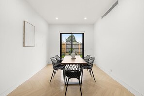 Un comedor con mesa de madera y sillas de estructura metálica negra, un cuadro en la pared y vistas a través de un ventanal grande.