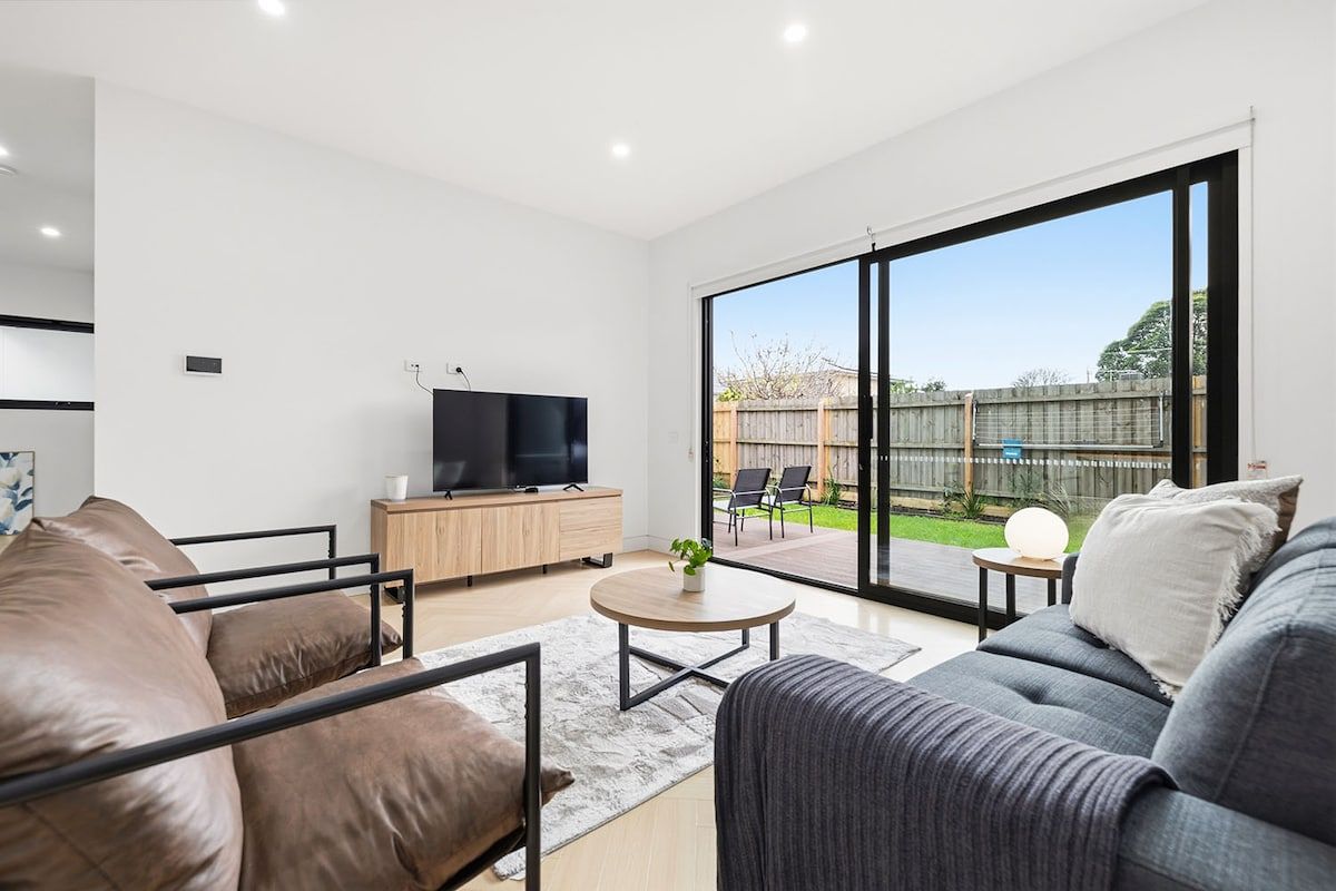 A modern living room with a flat-screen TV, leather sofas, a coffee table, and a large sliding glass door leading to an outdoor patio.