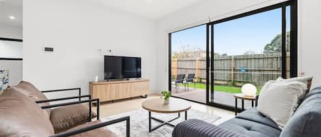 A modern living room with a flat-screen TV, leather sofas, a coffee table, and a large sliding glass door leading to an outdoor patio.