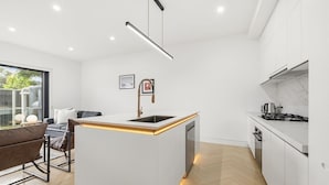 A modern kitchen with a central island, built-in appliances, and a dining area with a sofa and table. - Domi Rentals - The Cheltenham Southland House (Cheltenham)