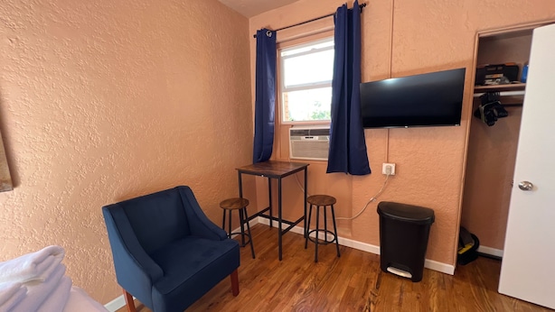 Ein Zimmer mit blauem Sessel, einem kleinen Tisch mit Hocker, einem an der Wand montierten Fernseher und einem Mülleimer.