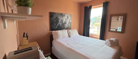 A hotel room with a bed, a nightstand, a mirror, a window with curtains, and a wall-mounted shelf with a plant.