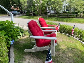 Ein roter Schaukelstuhl mit amerikanischer Flagge, platziert in einem Garten mit Holzpforte und viel Grün.