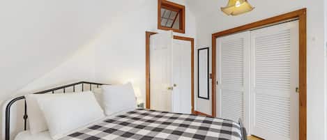 A bedroom with a checkered bedspread, a single bed with a headboard, a hanging light fixture, and a door with louvered shutters.