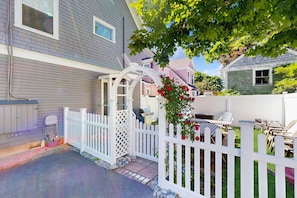A residential property with a white picket fence, a covered patio area, and a garden with red flowers. - Downtown Kennebunkport apartment with patio, steps from Dock Square (Kennebunkport)