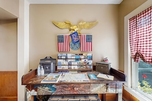 A room with a desk featuring a map, a small table with a book, a window with a checkered curtain, and a wall with an eagle decoration and flags. - 1 BR plus downtown - walk to Beaches/ Eateries (Kennebunkport)