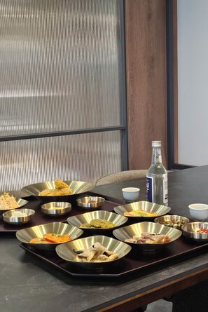 A dining table with multiple metal dishes containing various food items, a bottle of water, and a woven chair.