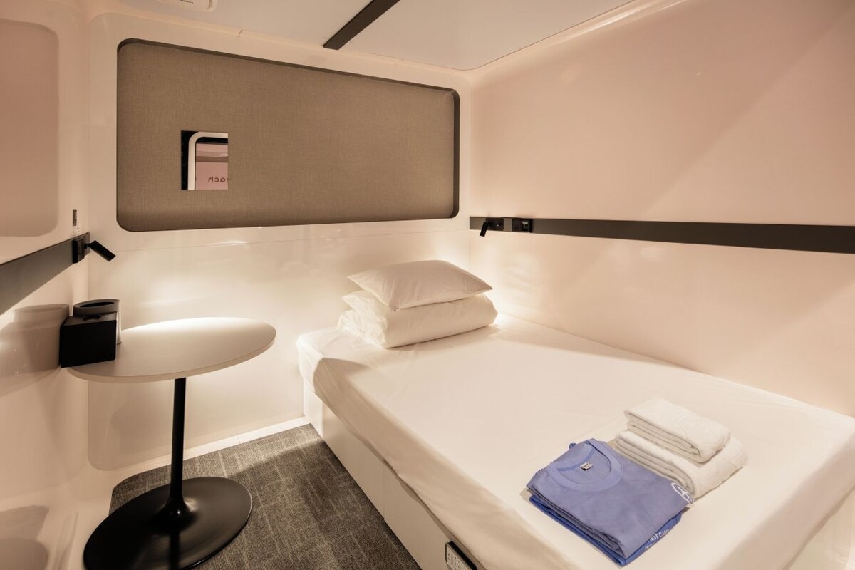 A compact hotel room with a bed, a small table, and a wall-mounted shelf.