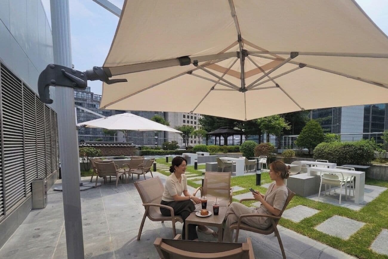 An outdoor seating area with tables and chairs under large umbrellas, surrounded by greenery and modern buildings.
