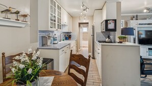 Une cuisine moderne avec des meubles blancs, un îlot central, un coin repas avec une table et des chaises en bois, et un téléviseur fixé au-dessus de l’îlot.