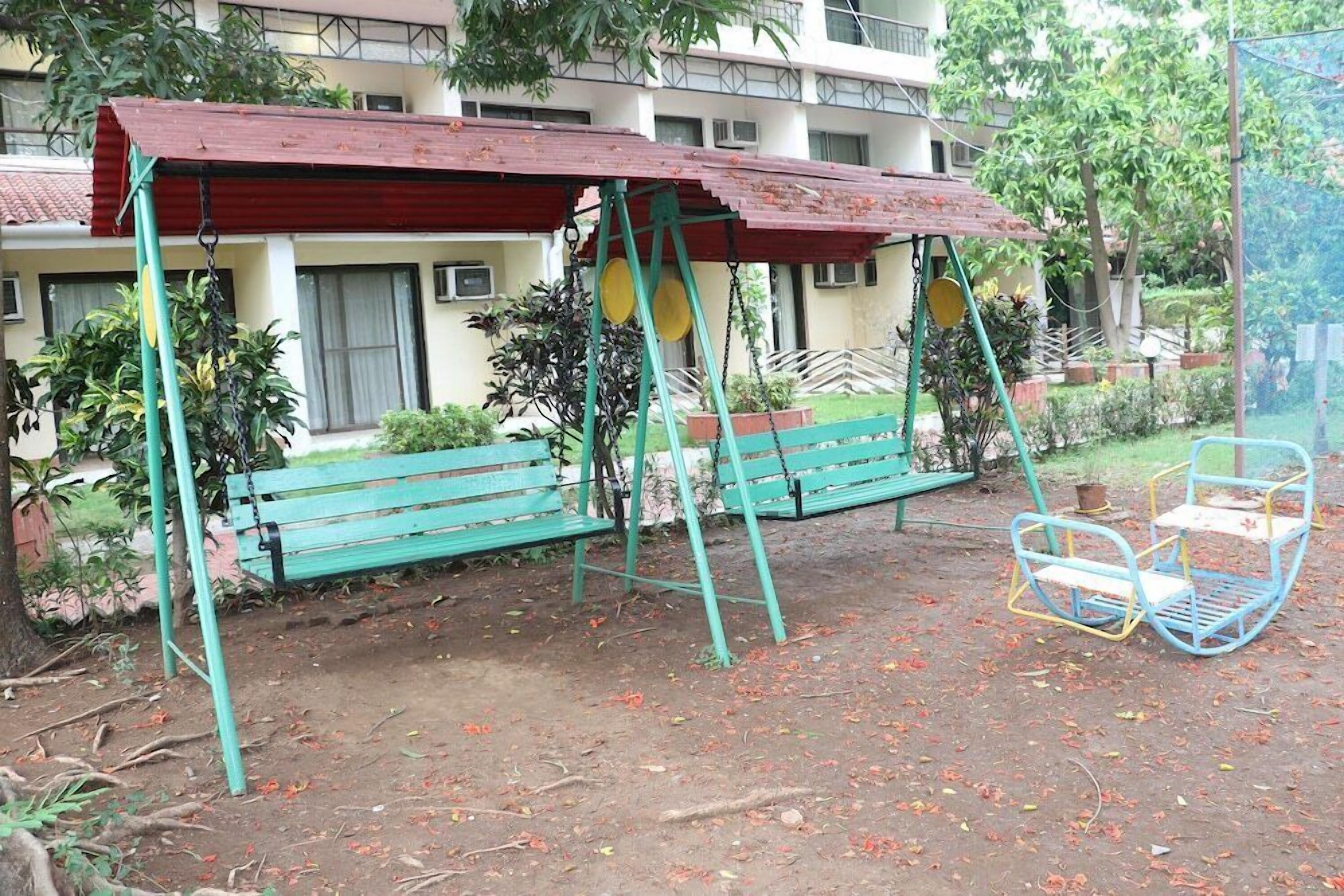 A playground with swings and a bench, surrounded by greenery and a building in the background.