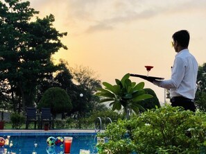 A person holding a tray with a drink and a ball, standing by a pool with a sunset in the background. - Vits Kamats Resort (Silvassa)