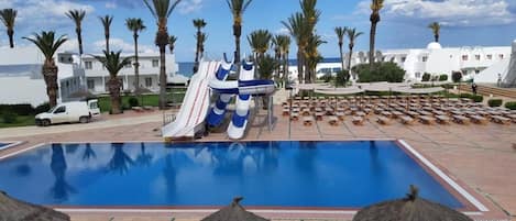En swimmingpool med vandrutsjebaner, liggestole, palmer og en bygning med hvid facade.