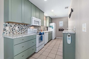 A kitchen with green cabinets, a white microwave, a white refrigerator, and a tiled backsplash.