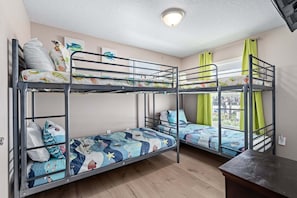 A bunk bed room with two beds, each with a colorful quilt, a window with curtains, and a wooden floor. - Ocean Views, Beach Across Street & Pet-Friendly | Sunrise Cottage (Flagler Beach)