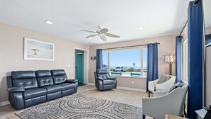 Ein Wohnzimmer mit einem schwarzen Ledersofa, einem gemusterten Teppich, einem grauen Sessel und Blick auf den Strand durch ein großes Fenster.