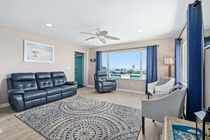 A living room with a black leather sofa, a patterned rug, a grey armchair, and a view of the beach through a large window. - Ocean Views, Beach Across Street & Pet-Friendly | Sunrise Cottage (Flagler Beach)