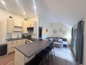 A modern kitchen with a central island, a dining area with bar stools, and a living space with a sofa and a coffee table. - Cabin - House of the Muse (Gordon)