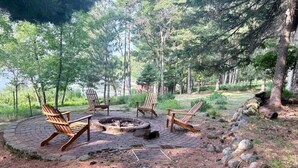 Ein runder Sitzbereich im Freien mit vier Adirondack-Stühlen um eine zentrale Feuerstelle, umgeben von einer natürlichen Waldlandschaft.