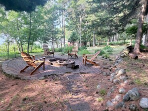 Ein runder Sitzbereich im Freien mit vier Adirondack-Stühlen um eine zentrale Feuerstelle, umgeben von einer natürlichen Waldlandschaft.