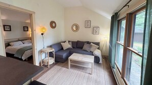 Ein gemütliches Wohnzimmer mit einer Couch, einem Couchtisch und einem Essbereich. An den Wänden hängen gerahmte Bilder und ein Fenster mit Vorhängen.