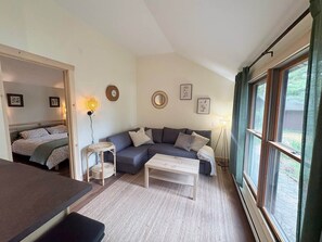 Ein gemütliches Wohnzimmer mit einer Couch, einem Couchtisch und einem Essbereich. An den Wänden hängen gerahmte Bilder und ein Fenster mit Vorhängen.