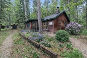 A log cabin with a paved driveway, surrounded by a garden with various plants and flowers. - Cabin - House of the Muse (Gordon)