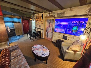 A room with a dining table, chairs, a sofa, a coffee table with papers, a television displaying a show, and a wall with a mural. - Wizard-Themed Condo Near Universal (Orlando)