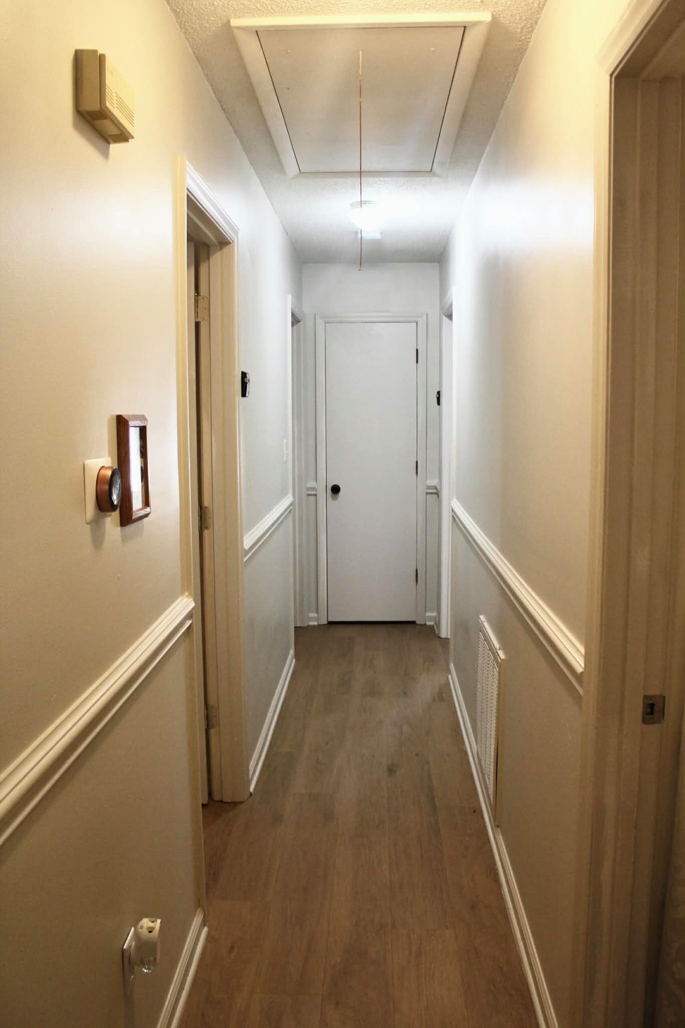 A narrow hallway with white walls and wooden flooring, leading to a closed door at the end.