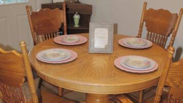 A round wooden dining table with six chairs, a framed welcome sign, and a vase on a side table.