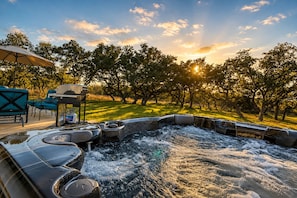 A hot tub with a surrounding deck, chairs, and a table, set against a backdrop of trees and a clear sky.