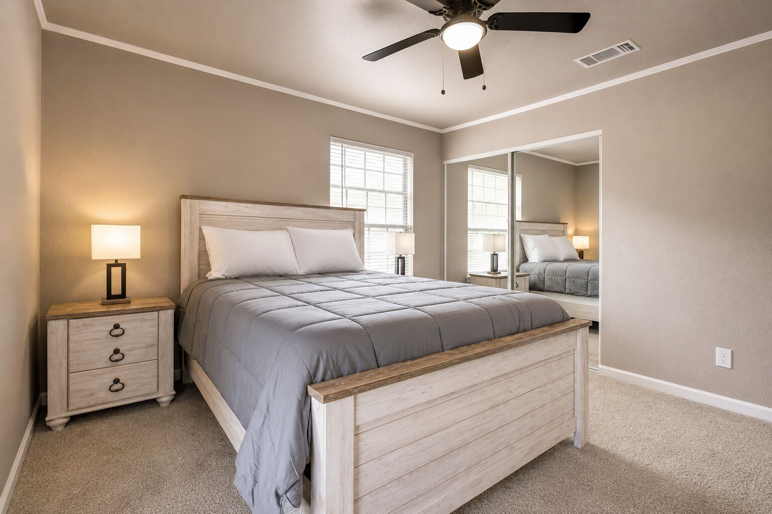 A bedroom with a bed, nightstand, lamp, ceiling fan, and a large mirror.
