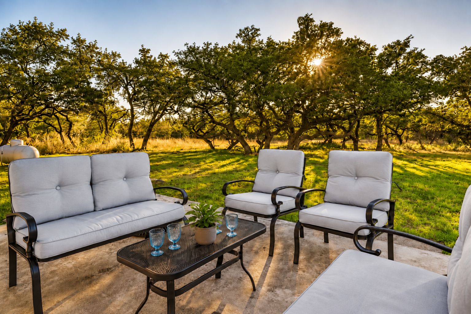 Ein Sitzbereich im Freien mit einer Couch und zwei Stühlen, einem kleinen Tisch mit Pflanze und Gläsern sowie Blick auf Bäume und Gras.