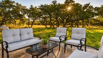 Ein Sitzbereich im Freien mit einer Couch und zwei Stühlen, einem kleinen Tisch mit Pflanze und Gläsern sowie Blick auf Bäume und Gras.