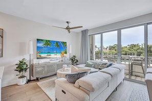 A modern living room with a large TV, a ceiling fan, a sofa, a side table, and a balcony with outdoor furniture. - Luxury Bayfront Designer Retreat • Paddleboard • Stunning Sarasota Sunsets (Sarasota)