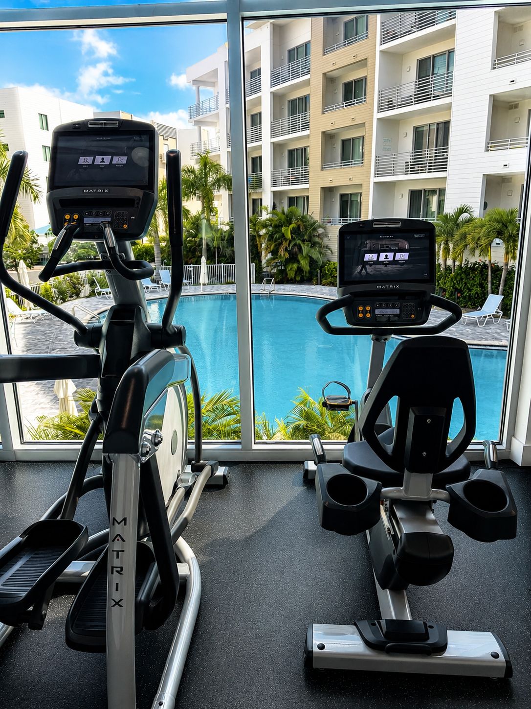 Un gimnasio con dos cintas de correr MATRIX, una piscina y vistas de un edificio de varios pisos con balcones.