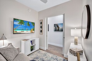Un salon moderne avec un téléviseur à écran plat, une console blanche, une lampe et un tableau représentant une scène de plage.