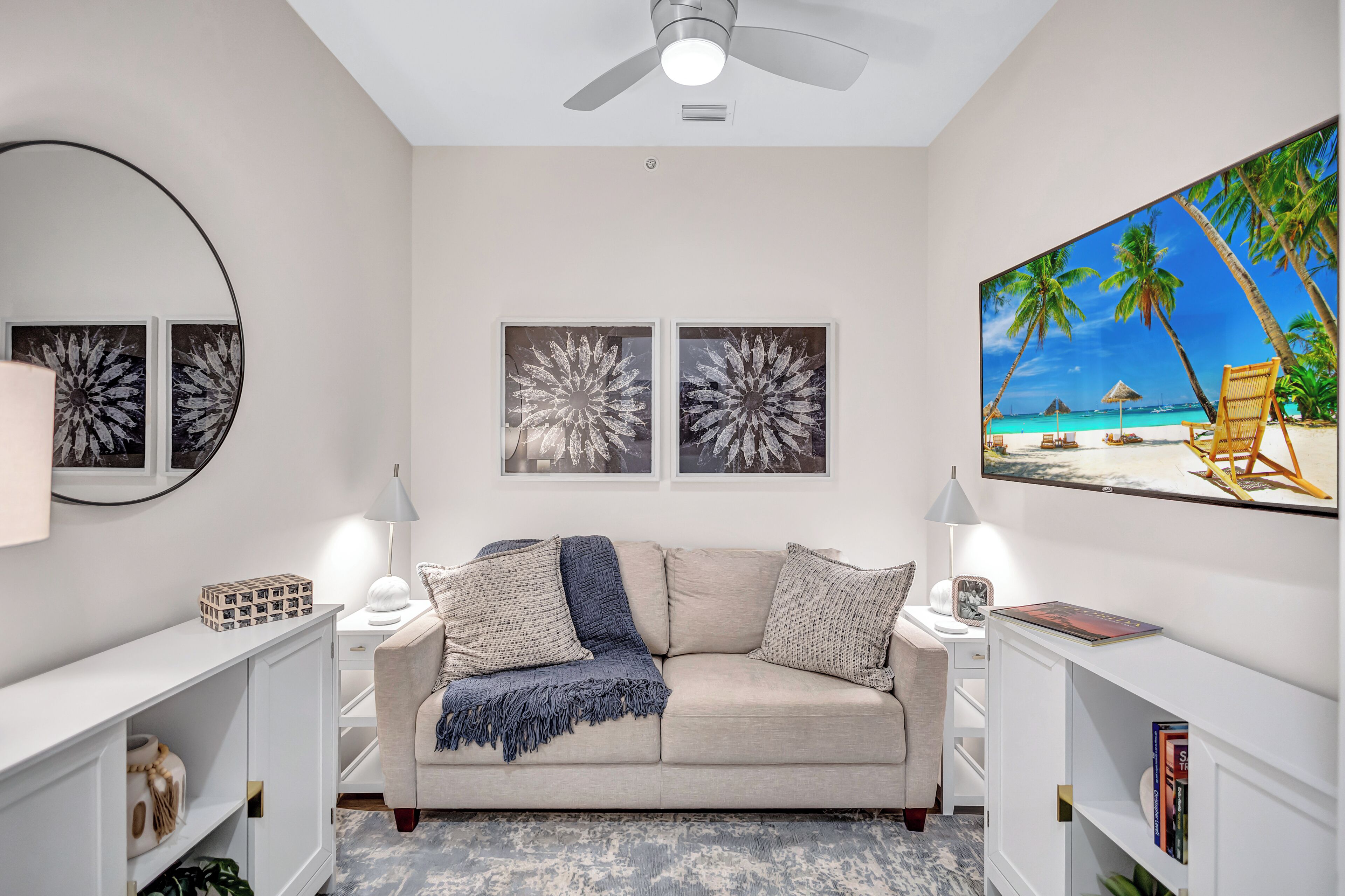 A living room with a beige sofa, two wall-mounted lamps, a ceiling fan, and a beach-themed painting.