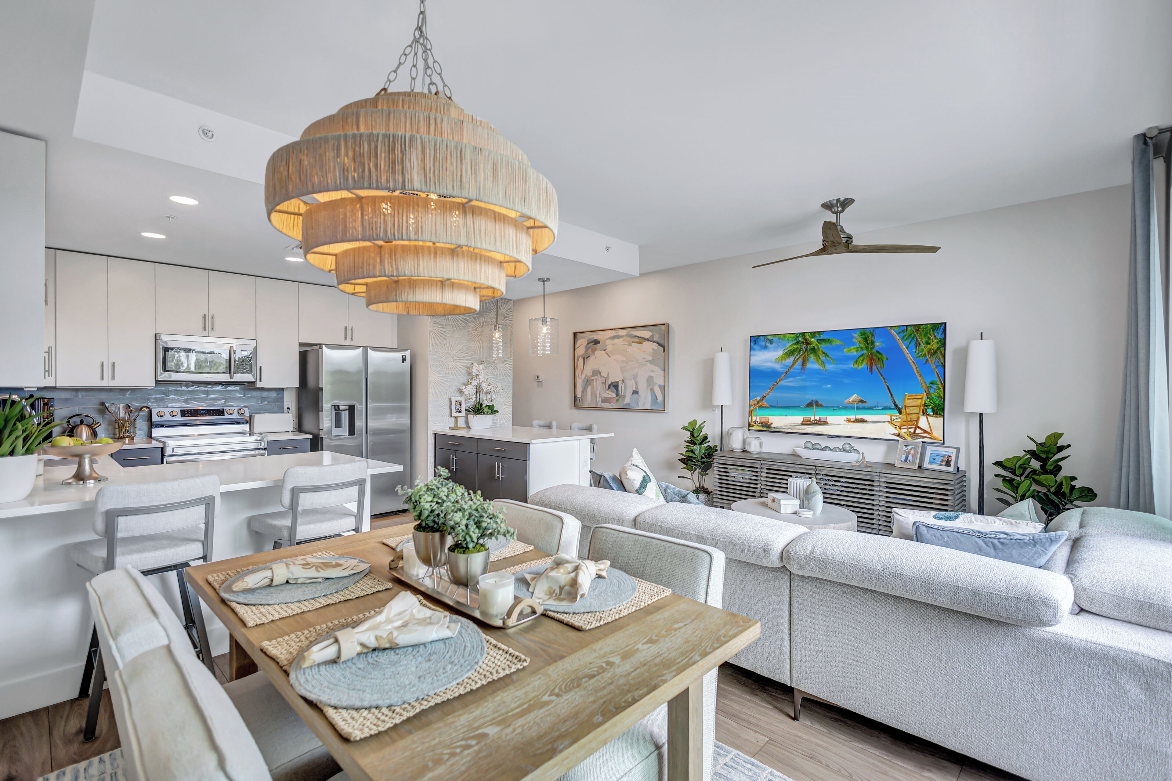 A modern kitchen with stainless steel appliances, a dining area with a wooden table, and a living room with a large sofa and a TV.