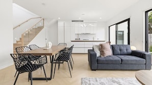 A modern living space with a dining area, sofa, staircase, kitchen, and large windows. - Domi Rentals - The Cheltenham Southland House (Cheltenham)