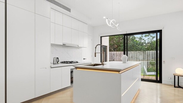 A modern kitchen with white cabinetry, a central island, and a large sliding glass door leading to a backyard. - Domi Rentals - The Cheltenham Southland House (Cheltenham)