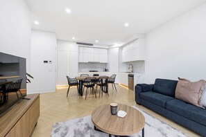 A modern living room with a sofa, coffee table, and dining area with chairs. - Domi Rentals - The Cheltenham Southland House (Cheltenham)