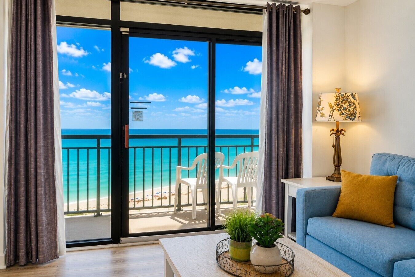 A room with a view of the beach, a blue sofa, a table with plants, and a lamp.