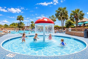 A pool area with a water play structure, lounge chairs, and palm trees. - Beach Cove Resort - Direct Oceanfront Balcony with Pools (North Myrtle Beach)