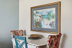 A dining area with a table, chairs, and a framed painting of a tropical scene. - Beach Cove Resort - Direct Oceanfront Balcony with Pools (North Myrtle Beach)
