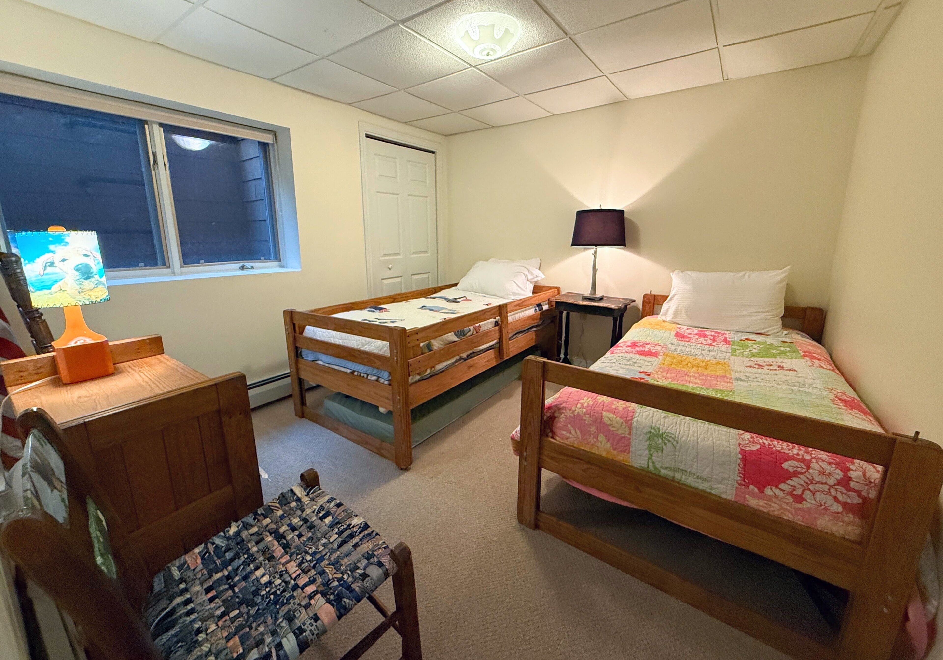 A room with two wooden beds, a desk with a lamp, a chair, and a window with blinds.